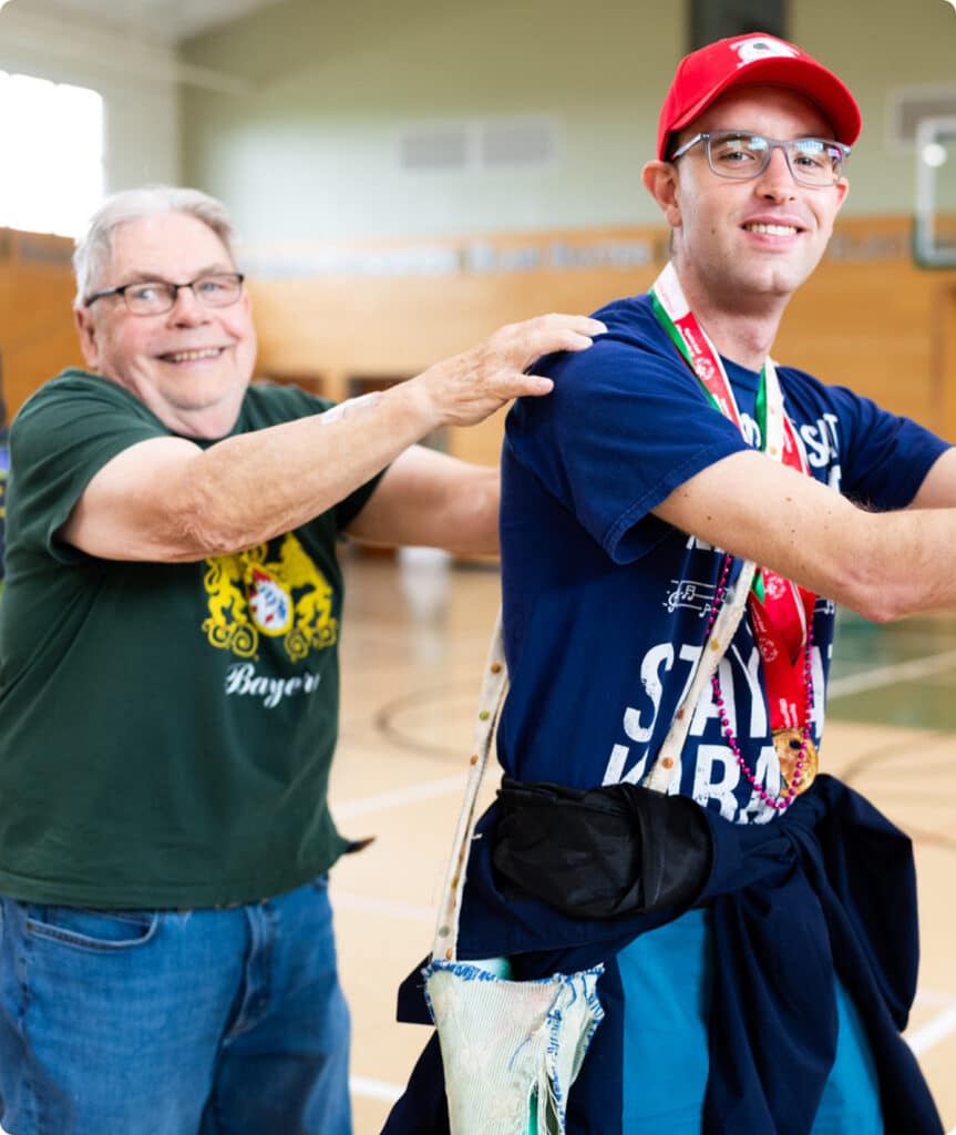 Two smiling men stand in a gym, one with his hands on the others shoulders, posing happily together.