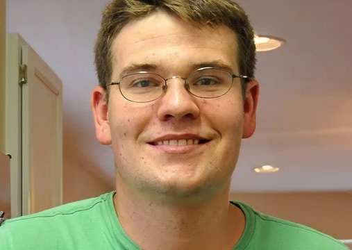 A man with short brown hair and glasses smiles, wearing a green shirt in a well-lit indoor setting.