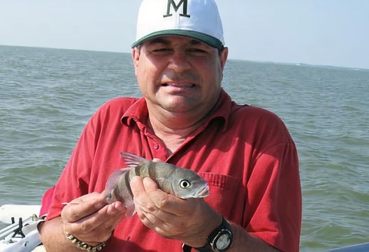 Man in red shirt and white cap holding a small fish, standing on a boat with water in the background.
