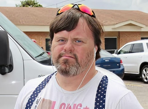 Man with a beard and sunglasses on his head, wearing earbuds and a white shirt, standing outside near cars.