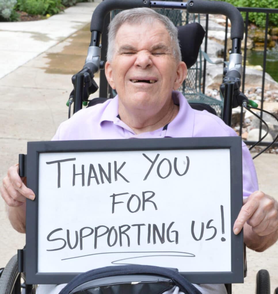 Smiling man in a wheelchair holds a sign that says, Thank you for supporting us!.