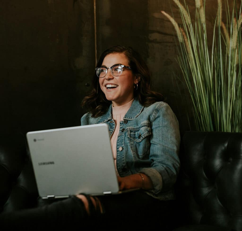 Woman wearing glasses and a denim jacket smiles while using a laptop on a dark sofa, with tall plants nearby.