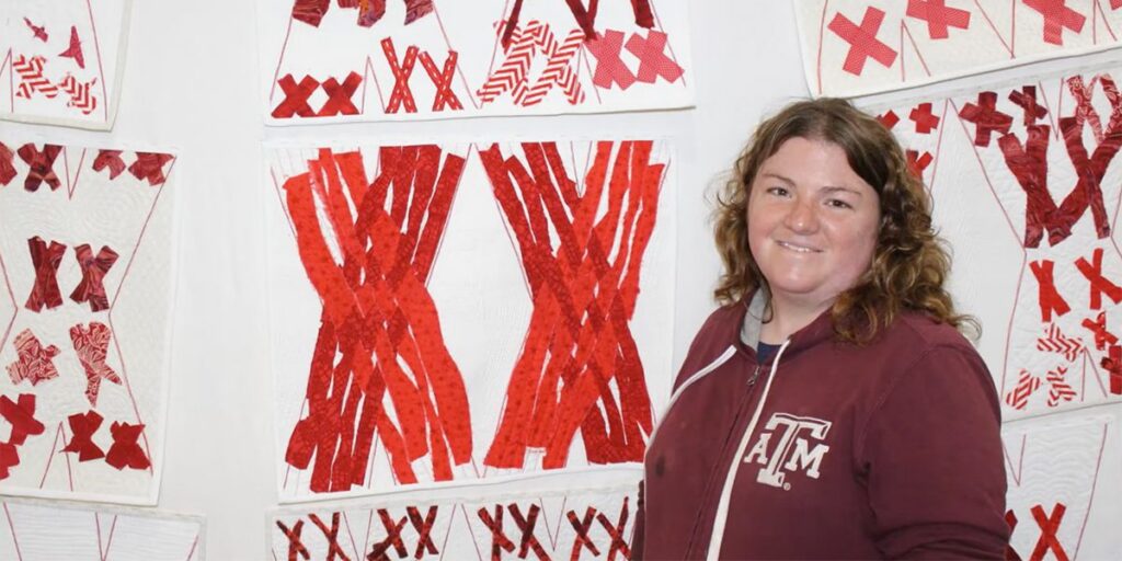 A woman in a Texas A&M hoodie stands by quilt panels with red X patterns on a white background.