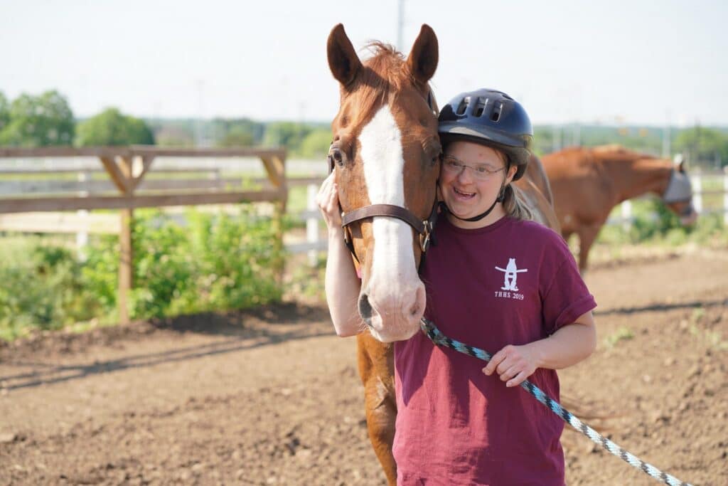 Smiling woman in a helmet hugs a brown horse on a sunny day at an outdoor riding area.