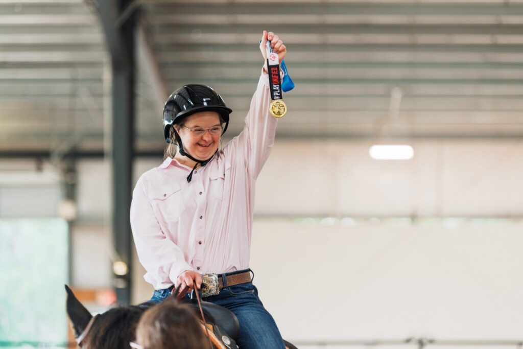 A smiling person in a helmet on horseback proudly holds up a gold medal inside an indoor arena.