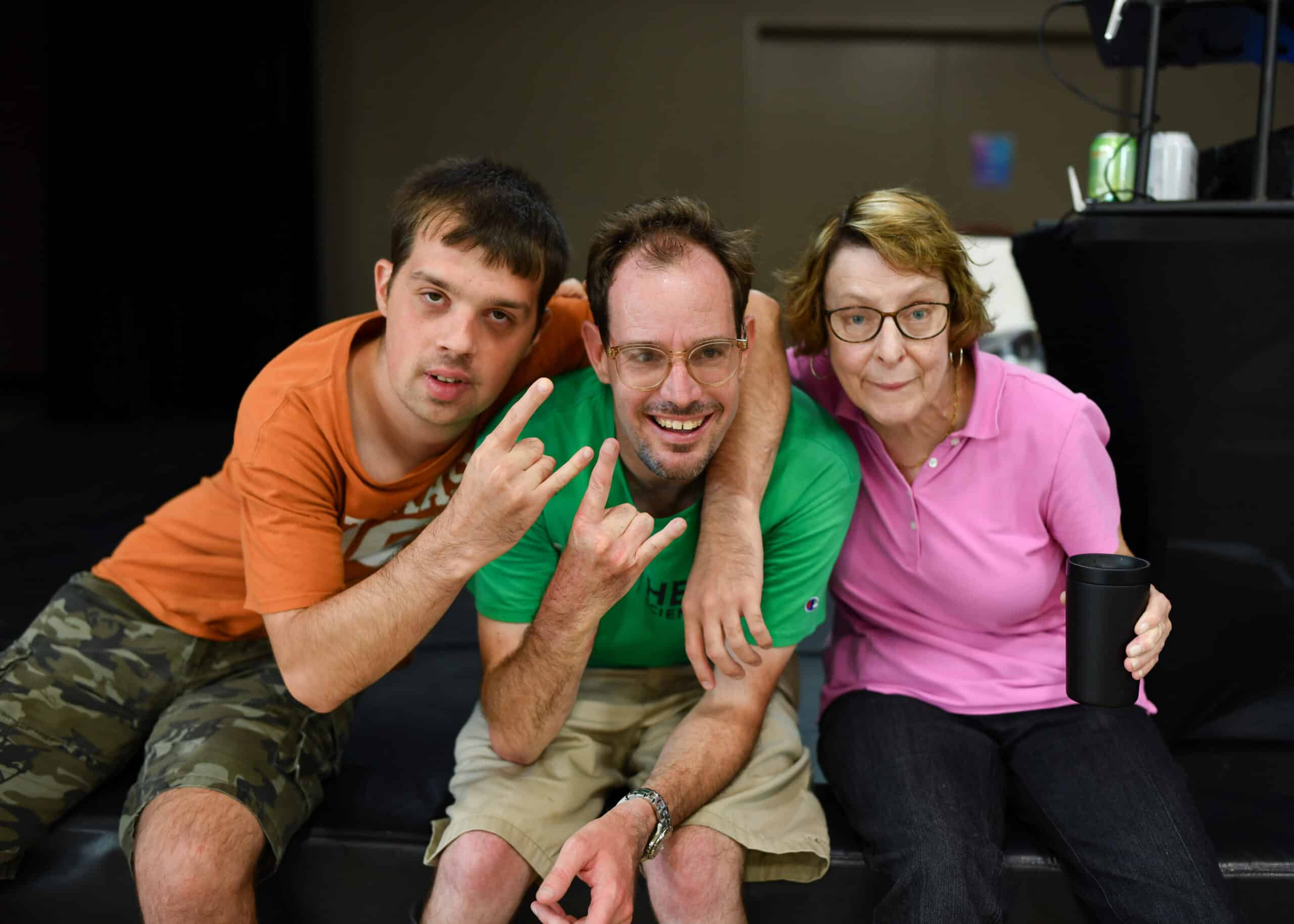 Three people sitting together, smiling; one making a hand gesture, another holding a cup, all looking at the camera.