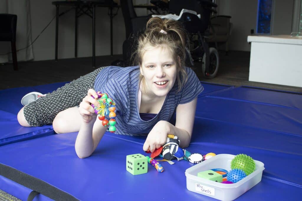 A smiling girl lies on a blue mat, playing with colorful sensory toys spread out in front of her.