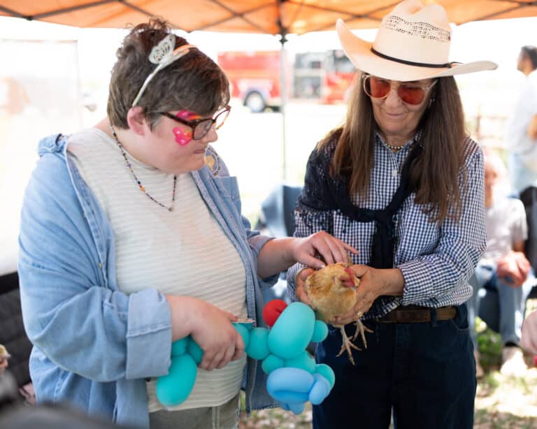 Two women pet a chicken under a tent; one holds blue balloon animals, the other wears a cowboy hat and sunglasses.