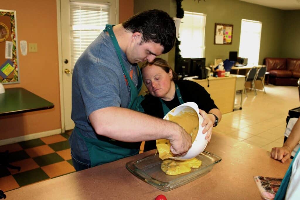 Two people pour yellow cake batter from a bowl into a baking dish in a kitchen setting.