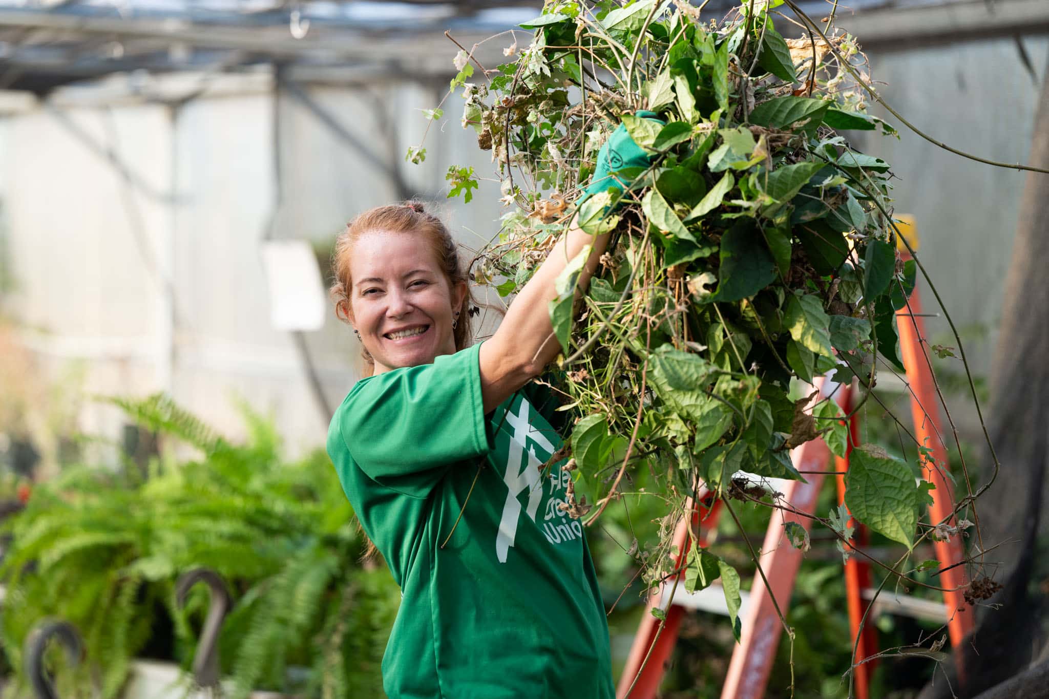 Smiling woman in a green shirt lifts a pile of garden plants and leaves inside a greenhouse.