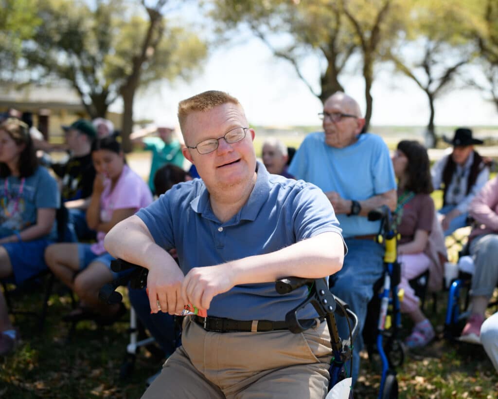 Smiling man sitting outdoors among a group of people at a sunny event.