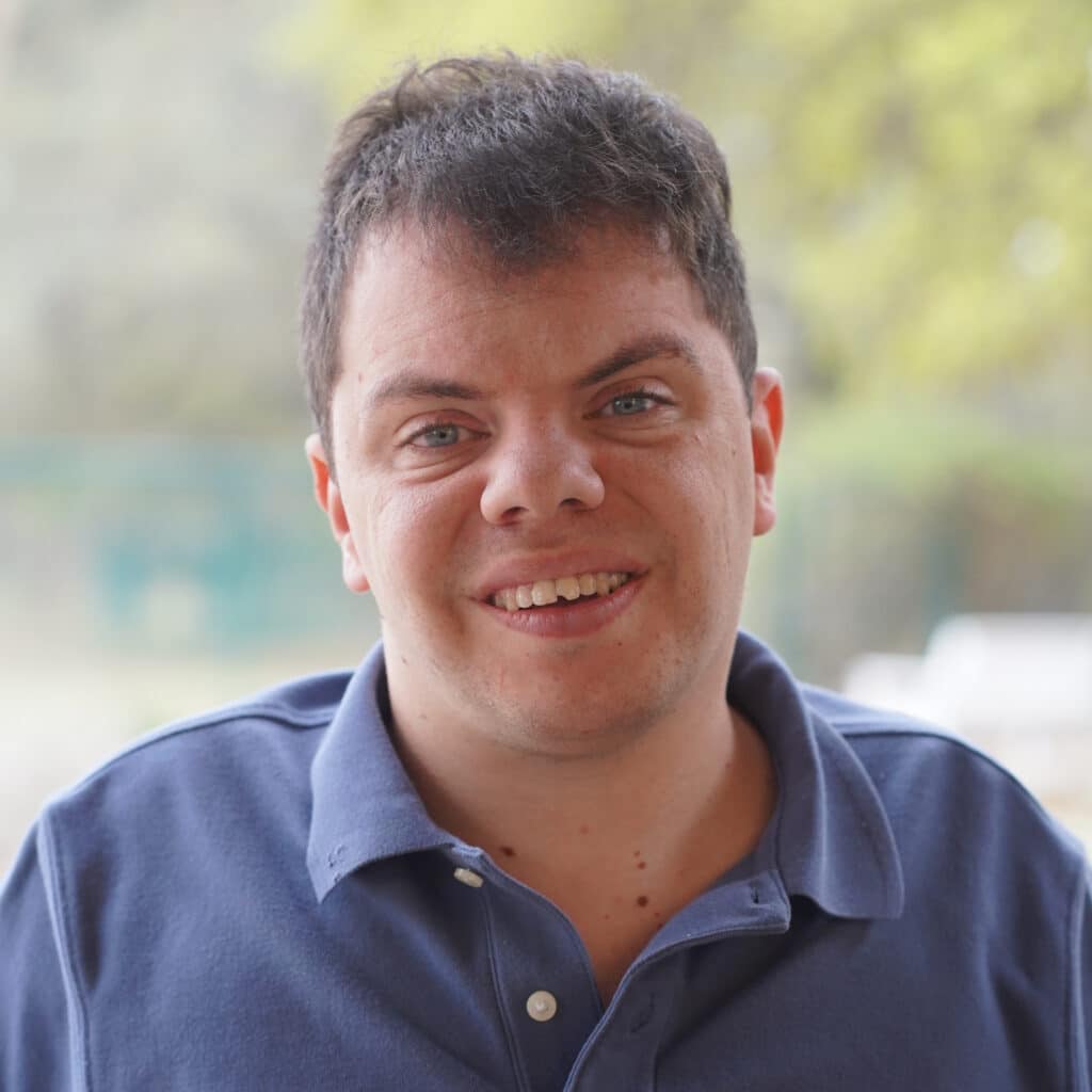 Man with short dark hair smiling, wearing a blue polo shirt, with a blurry outdoor background.