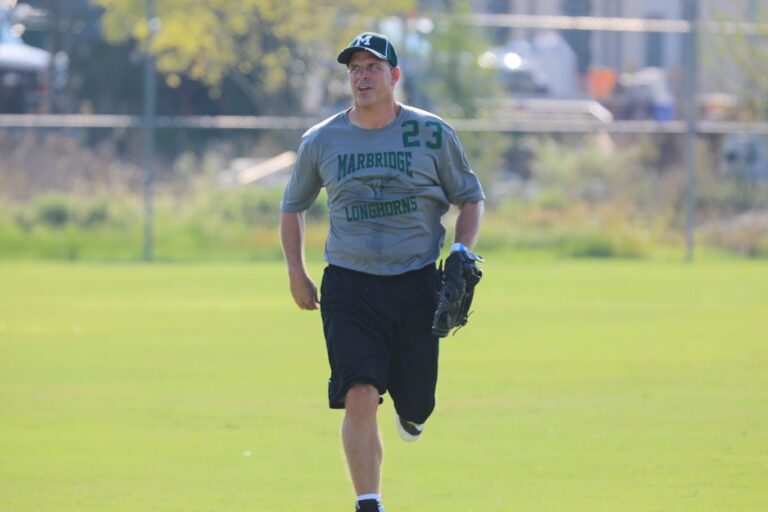 Man in a gray Marbridge Longhorns shirt and cap running on a grassy field with a baseball glove.