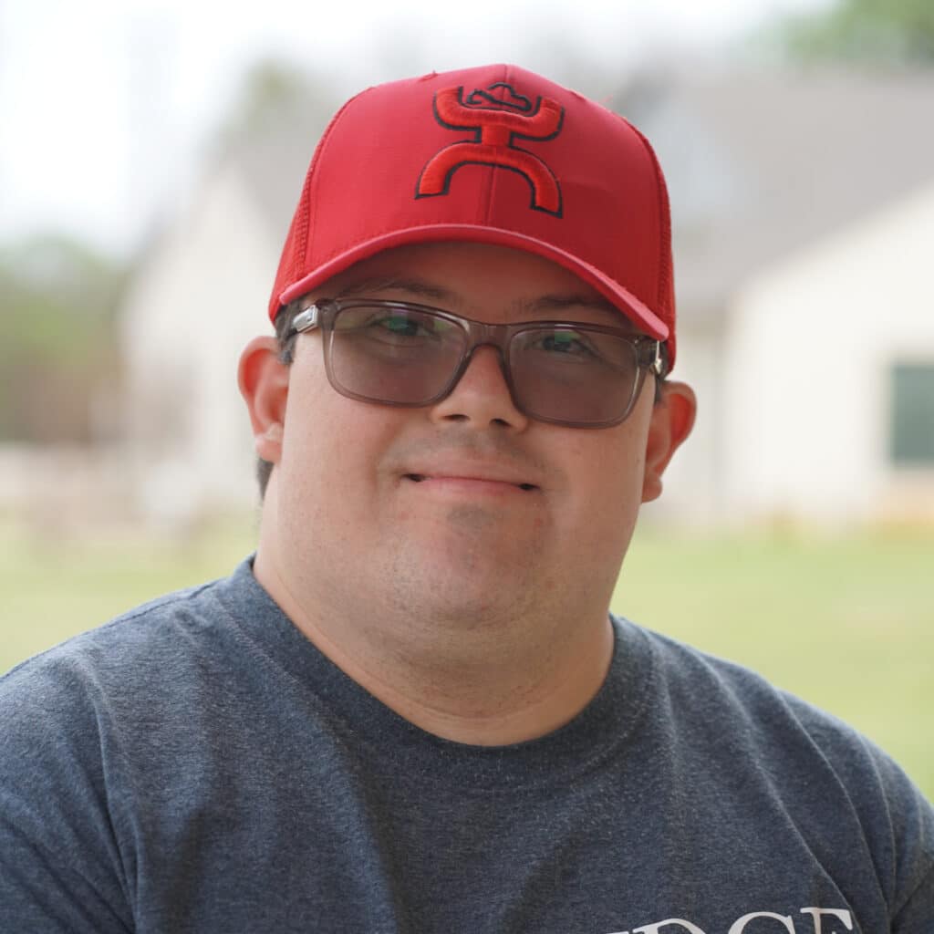 A person wearing glasses and a red cap, smiling outdoors with blurred buildings in the background.