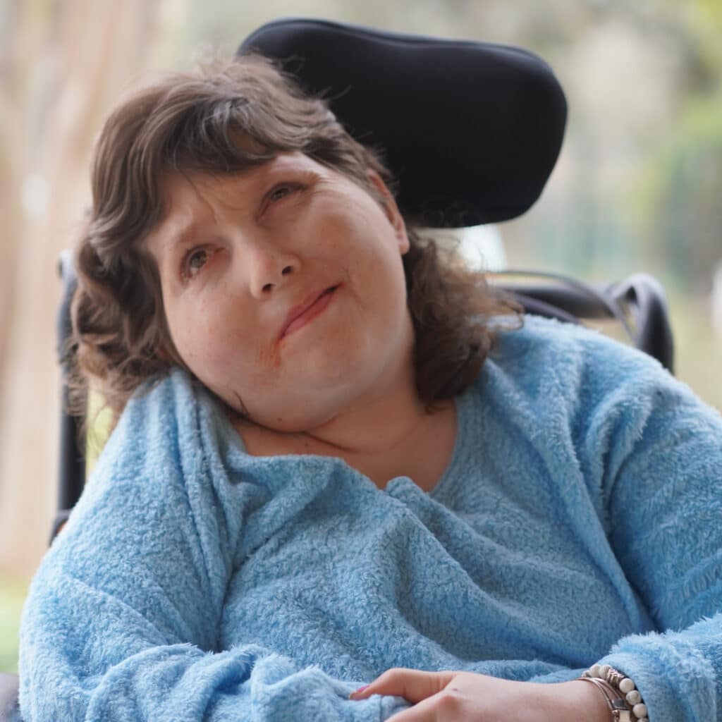 Woman in a wheelchair wearing a light blue fleece, looking up with a gentle expression.
