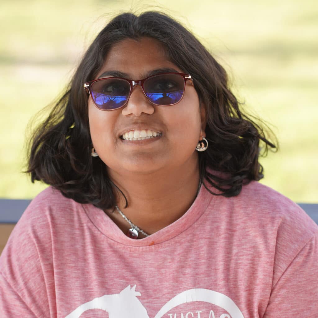 Smiling person with shoulder-length dark hair, sunglasses, and a pink shirt, outdoors on a sunny day.