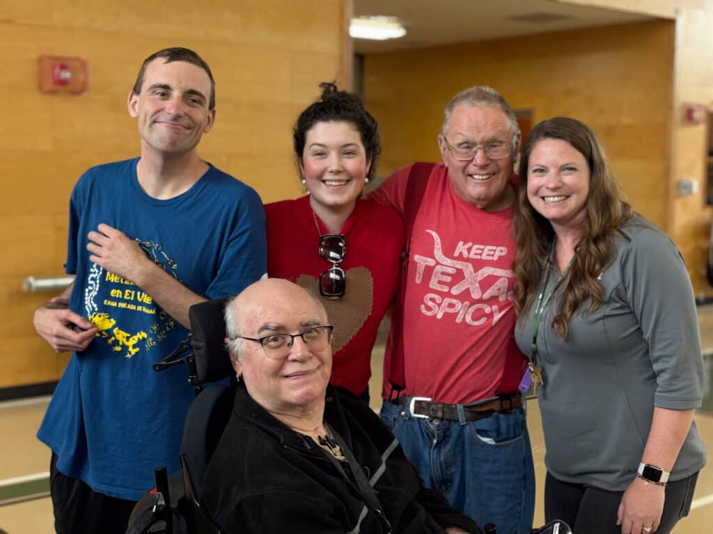 Five smiling people, including one in a wheelchair, pose together indoors against a wooden wall.