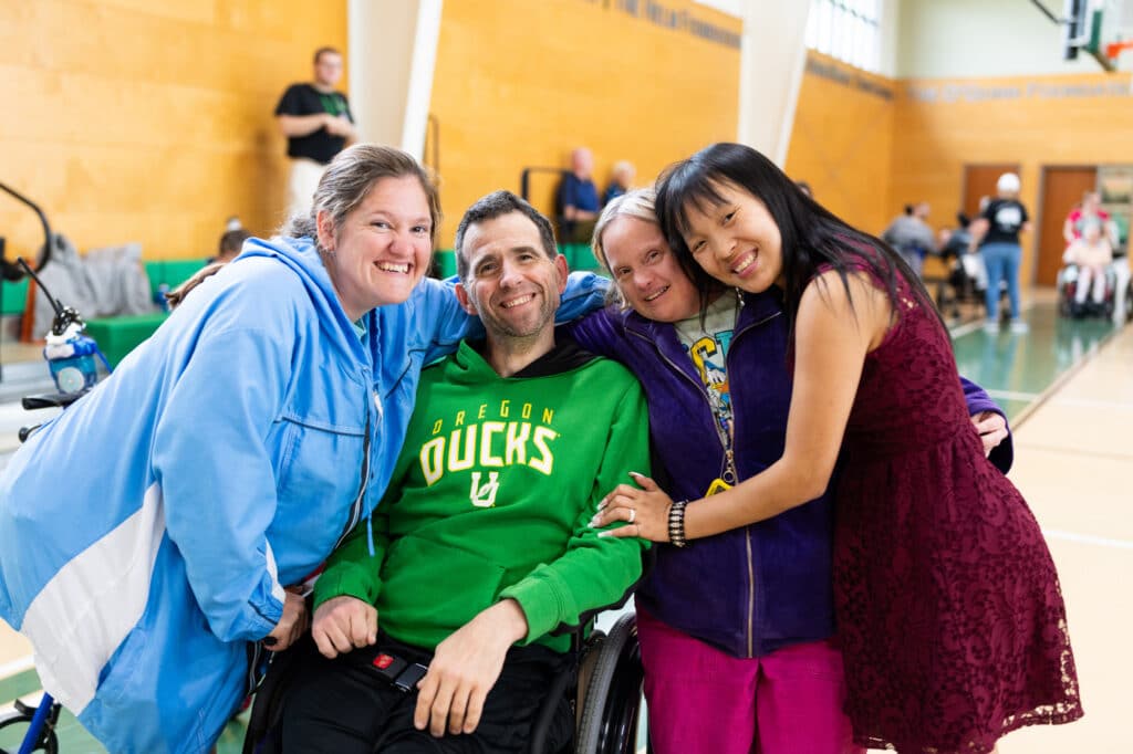 Four smiling friends, one in a wheelchair, pose together in a gymnasium, showing warmth and camaraderie.