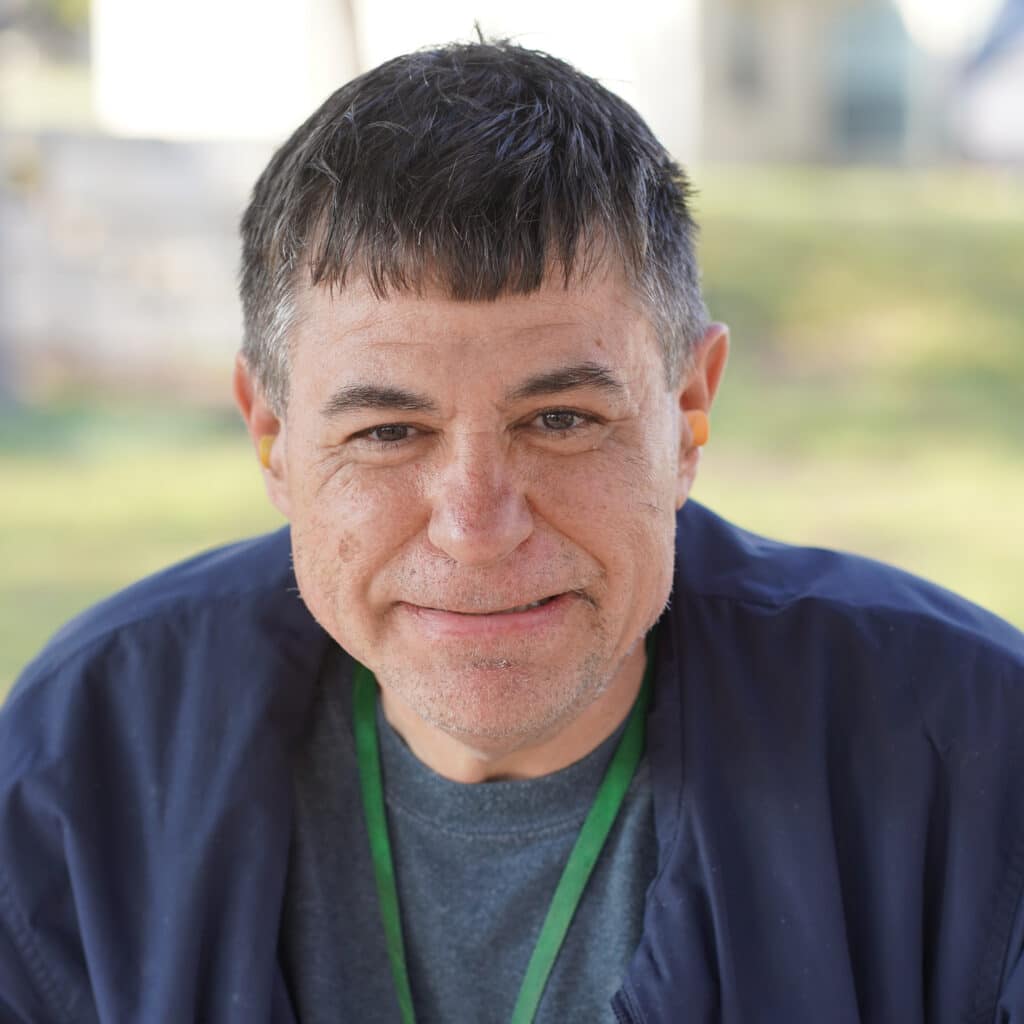 A man with short dark hair and a navy jacket smiles at the camera outdoors.