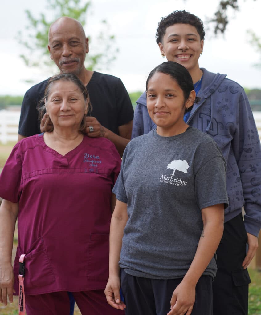 Four people stand outdoors, smiling, two in scrubs and two in casual clothes, with trees in the background.