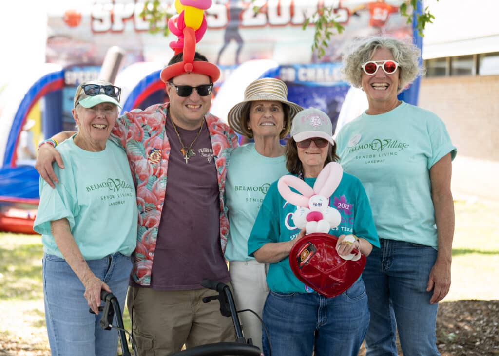 Five smiling adults pose outdoors at an event, some wearing teal shirts and festive hats, with inflatables behind them.