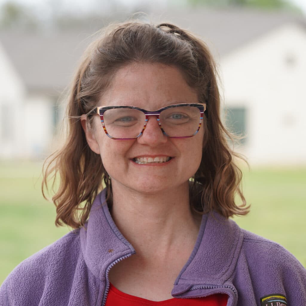 Woman with brown hair and glasses smiling, wearing a purple fleece jacket, outdoors with houses blurred behind.
