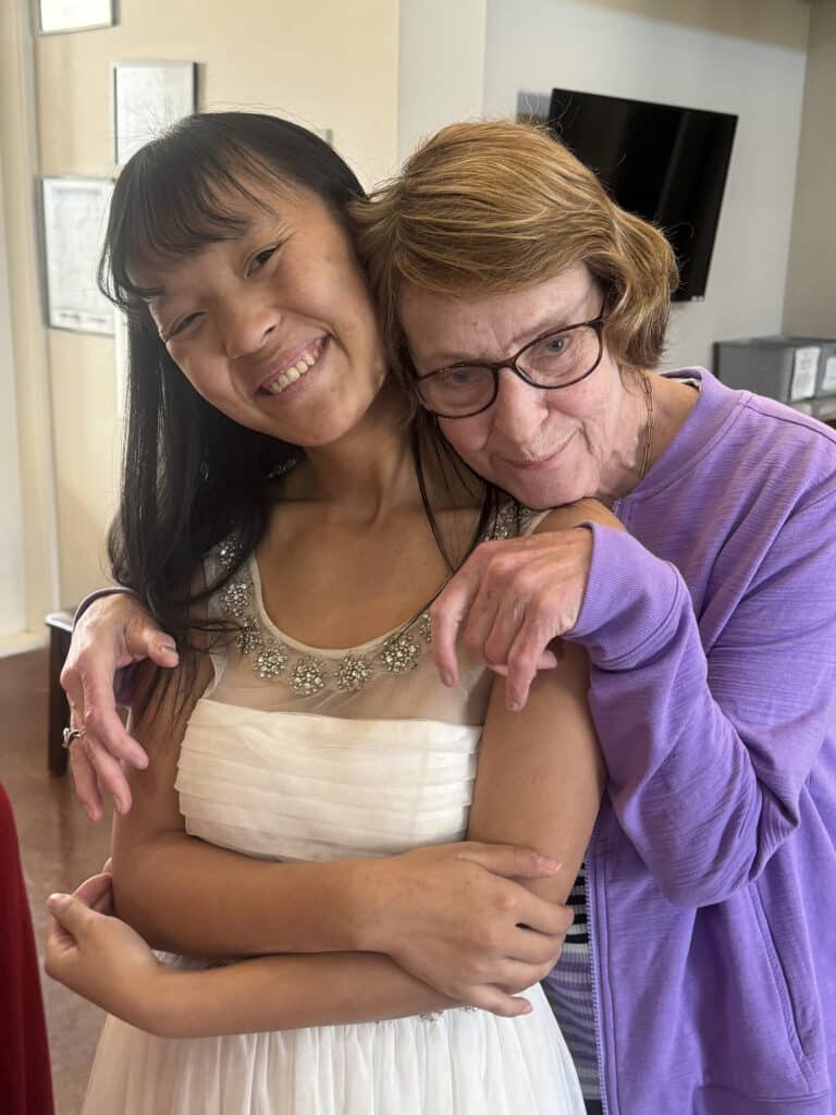 An elderly woman hugs a smiling girl in a white dress; both are happy and facing the camera indoors.