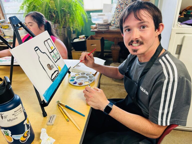 A man smiles while painting a bottle and glass at a table in an art classroom, wearing a black apron.