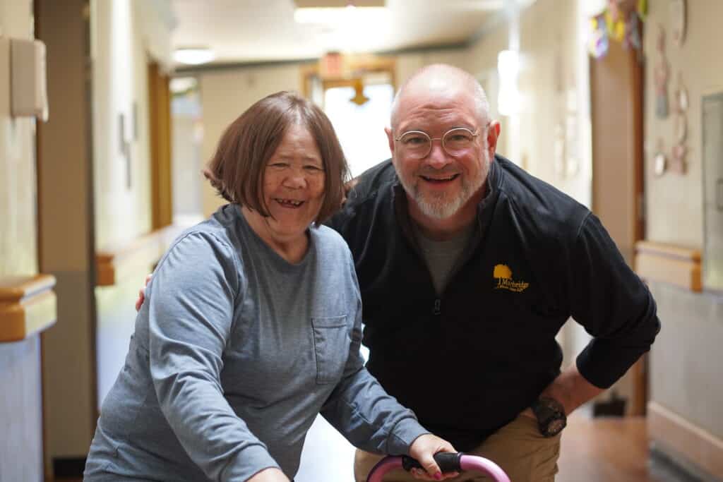 Smiling woman with a walker and a man posing together in a brightly lit hallway.
