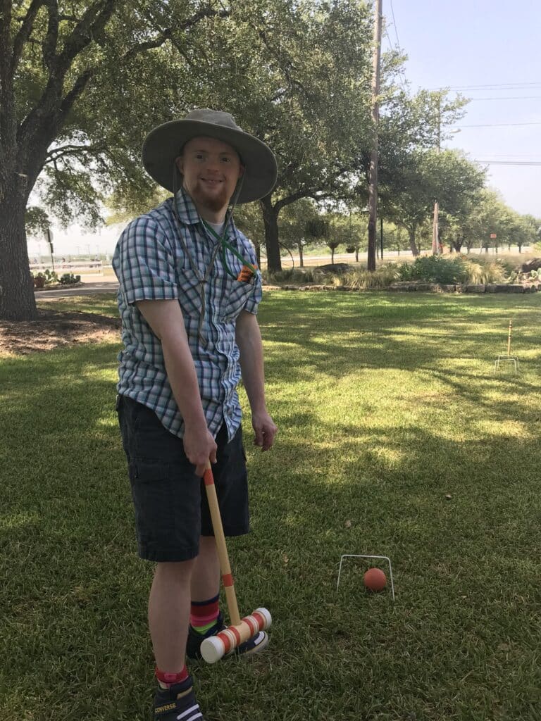 A man in a hat smiles while playing croquet on a grassy lawn with trees in the background.