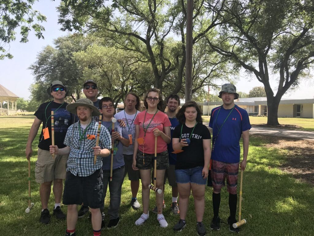 A group of nine people posing outdoors on grass, holding croquet mallets, with trees and buildings in the background.