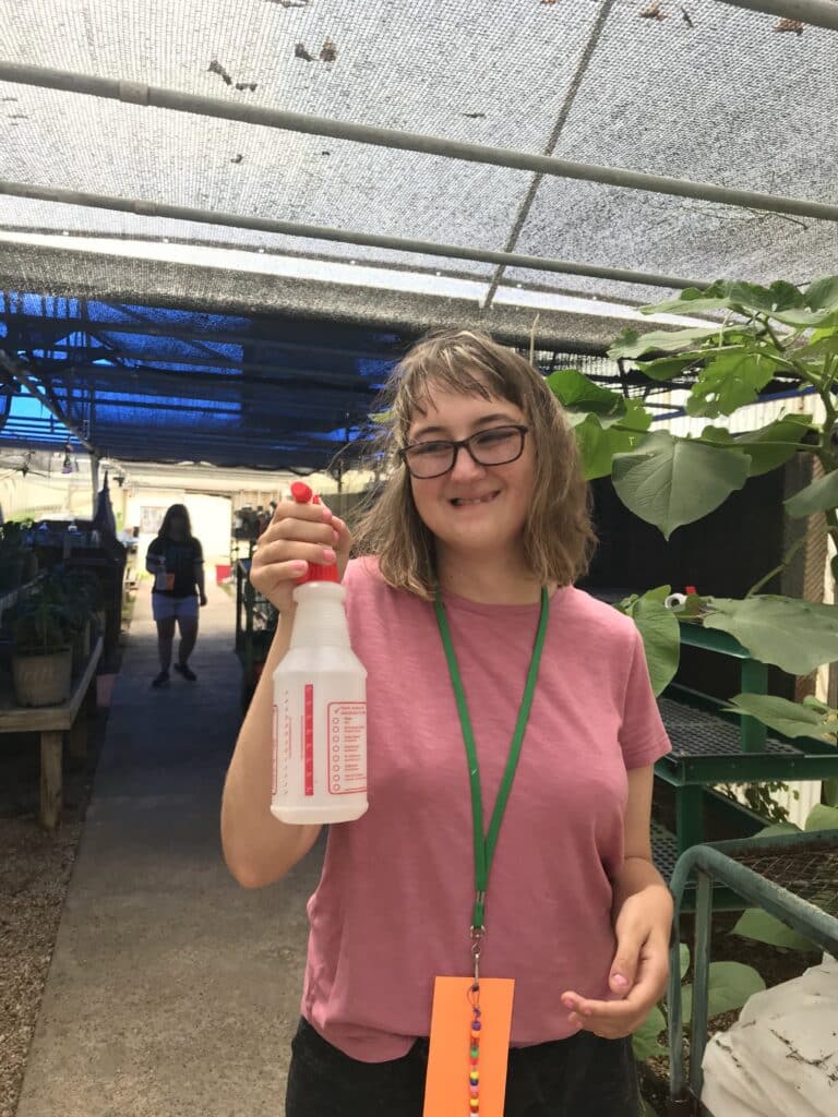 A smiling woman in a pink shirt holds a spray bottle in a greenhouse with plants around her.