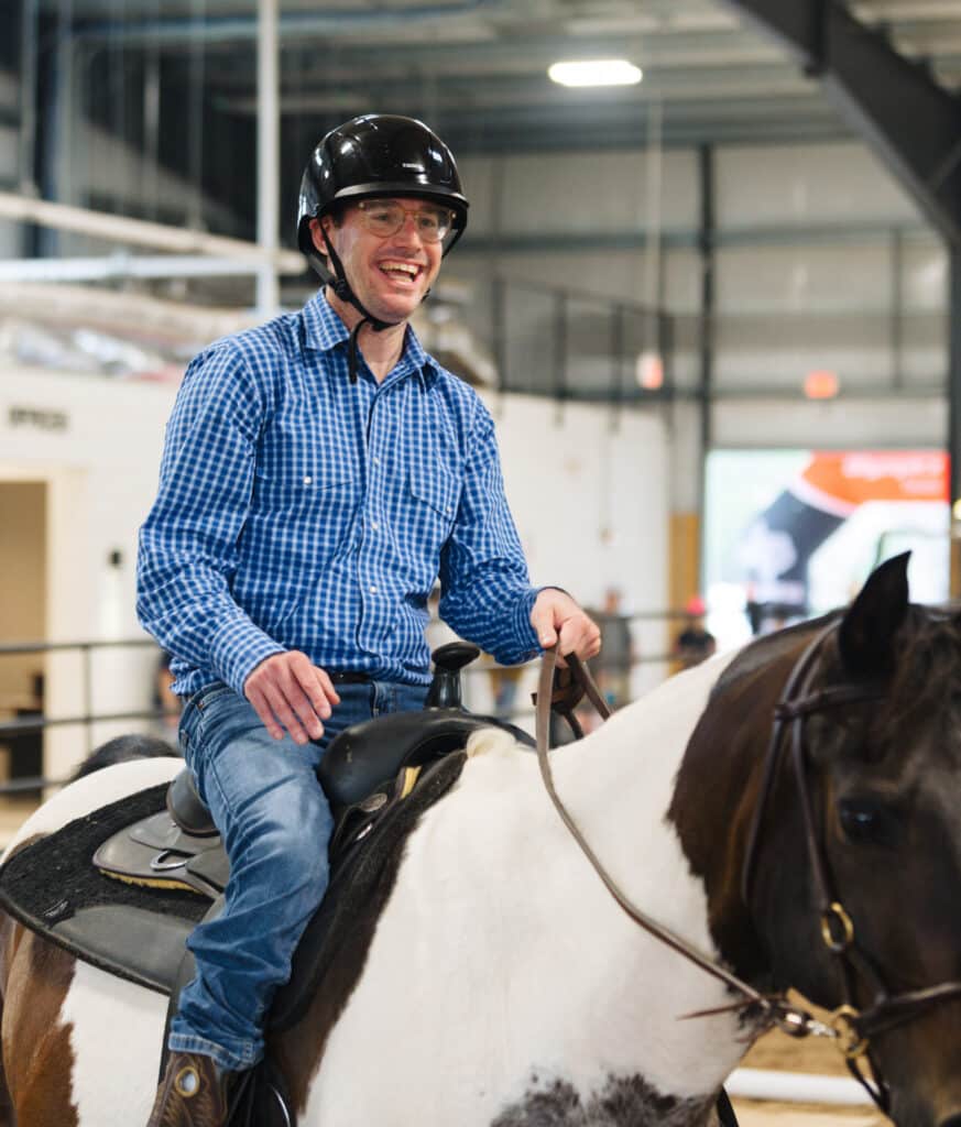 A smiling man in a blue plaid shirt and helmet rides a black and white horse indoors.
