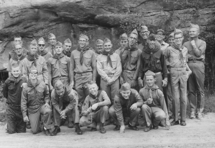 A Group Of Boys And Men In Scout Uniforms Pose In Front Of A Rock Formation Outdoors.