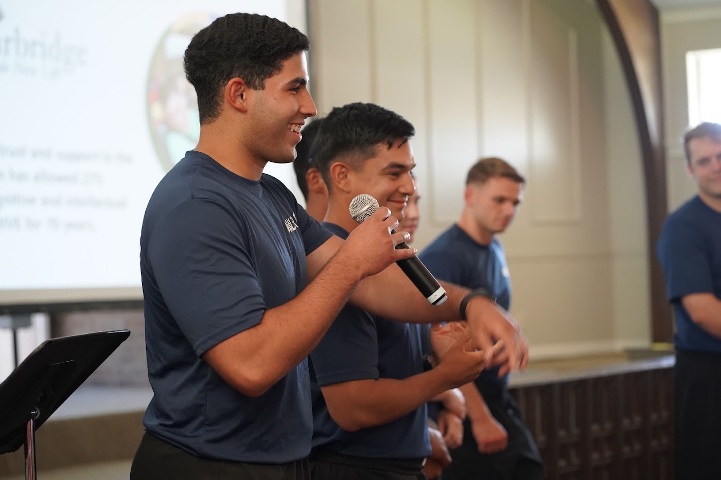 A group of young men wearing matching blue APD shirts stand side by side, with one speaking into a microphone and the others smiling.