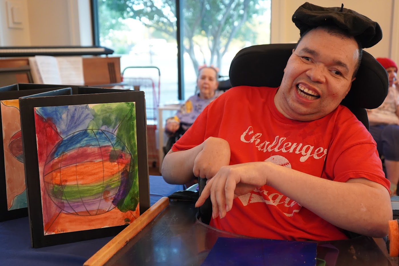 A man in a red shirt, sitting in a wheelchair, smiles next to colorful paintings displayed on a table.