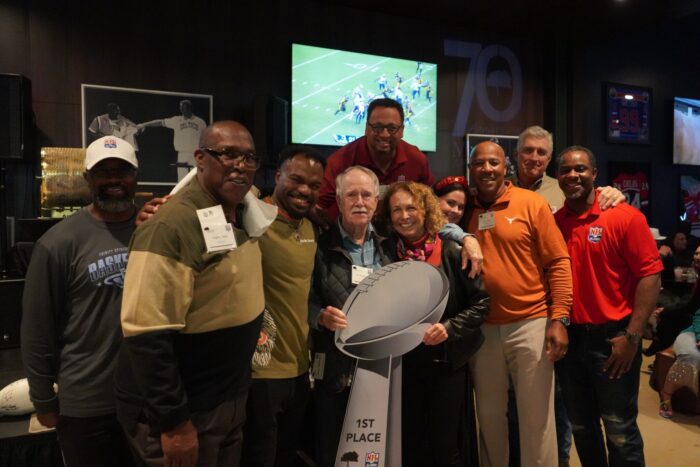 A Group Of People, Including A Man And Woman Holding A 1st Place Trophy, Pose For A Photo In A Sports-themed Room.