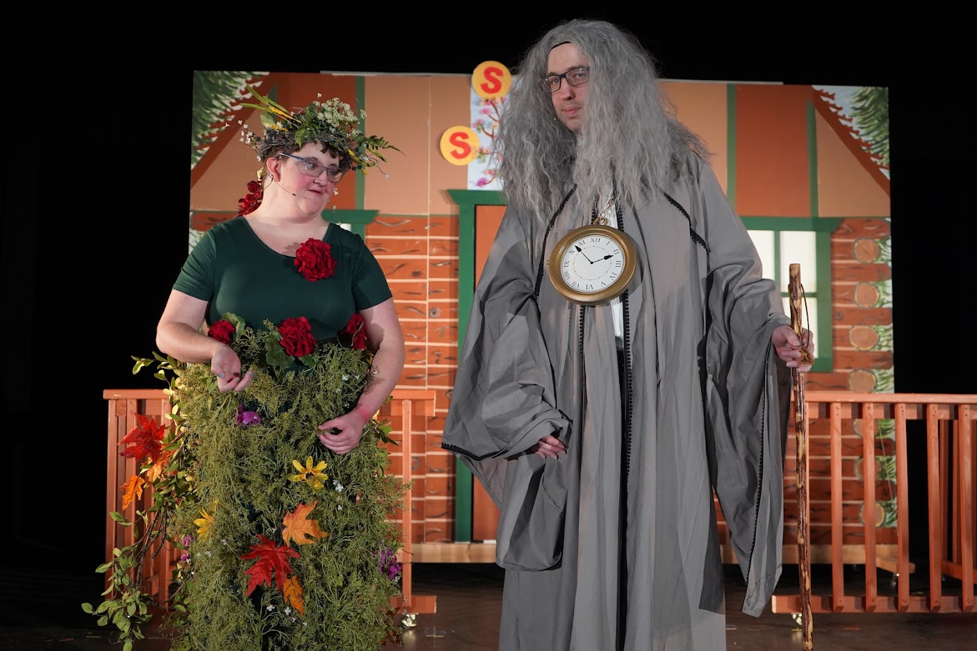 Two people in theatrical costumes: one dressed as a tree with flowers and greenery, and the other as an old figure with a gray wig, a clock pendant, and a staff.