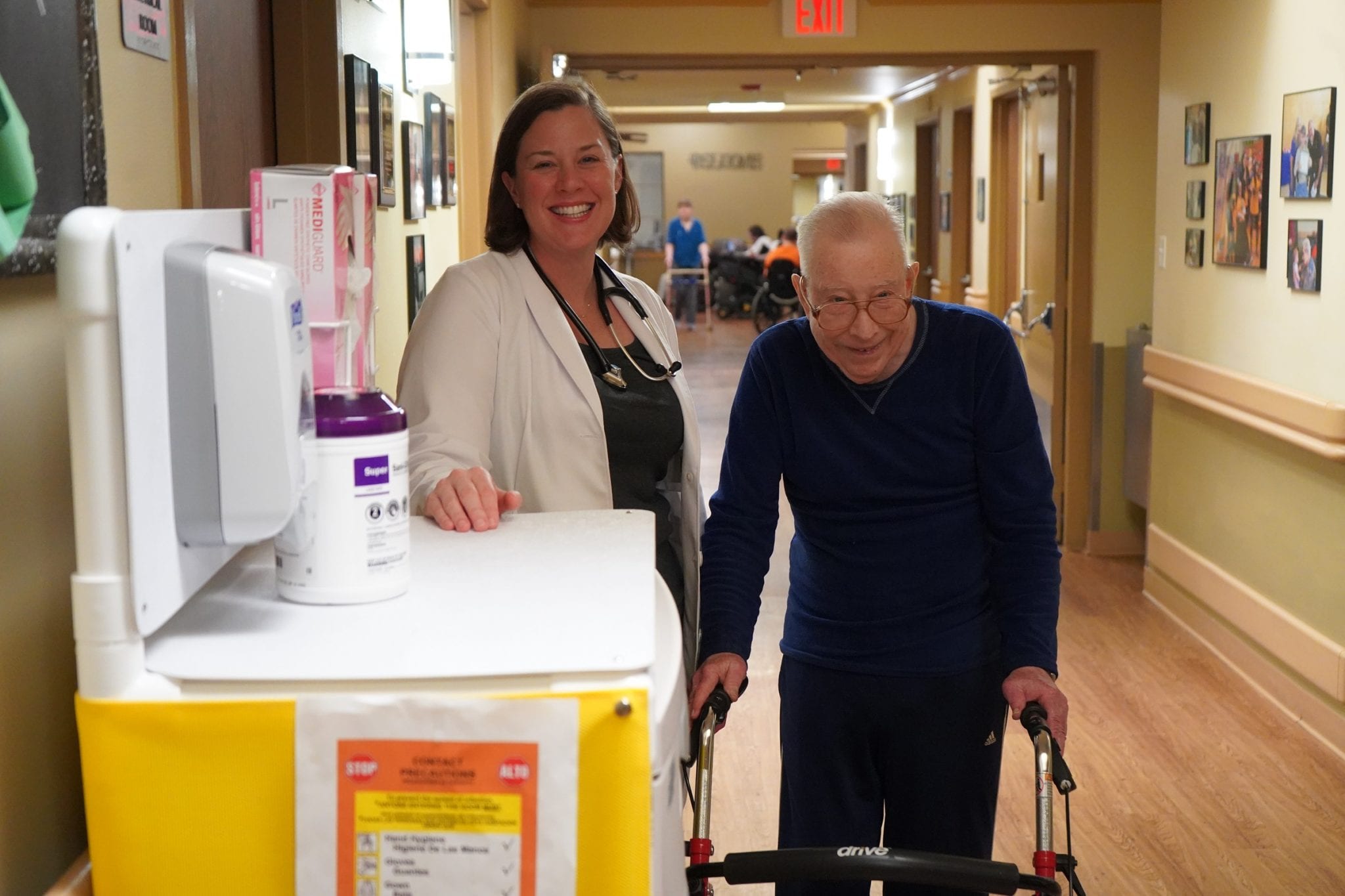 A mature male resident smiles and standing next to a smiling female healthcare worker in a corridor.