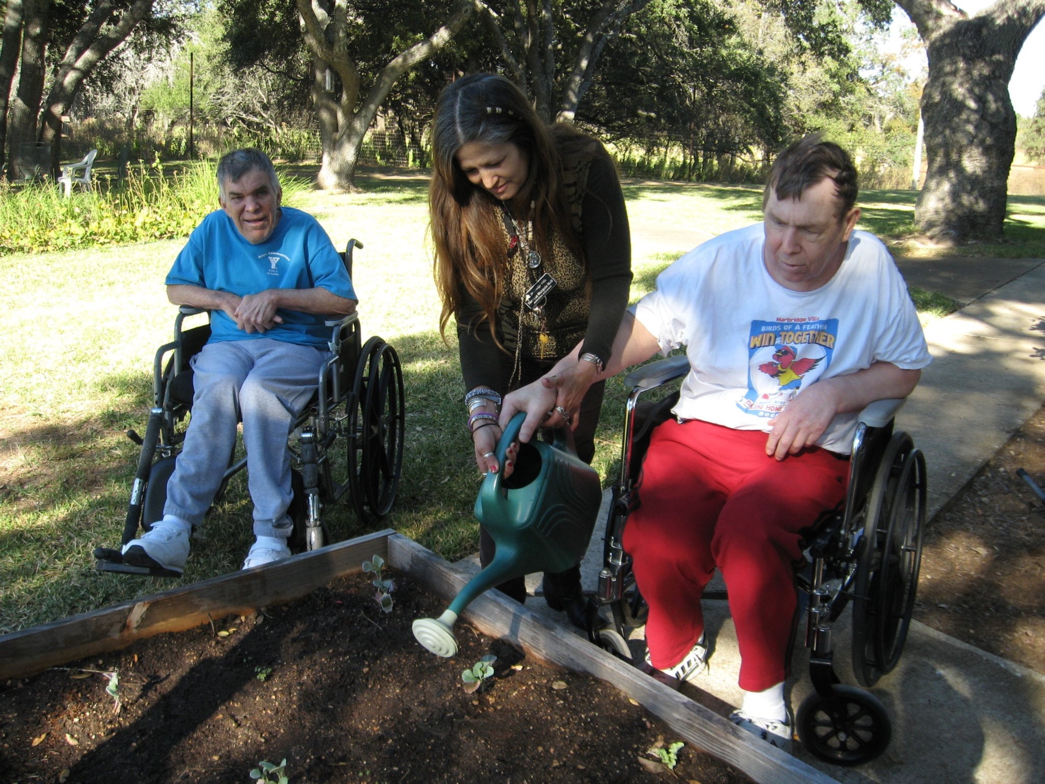 A woman helping a man in a wheelchair water plants in a raised bed garden.