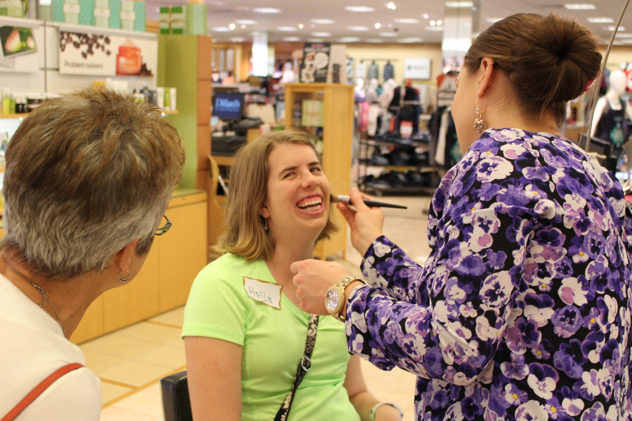 A smiling female resident has makeup applied by a beautician at a department store cosmetics counter.