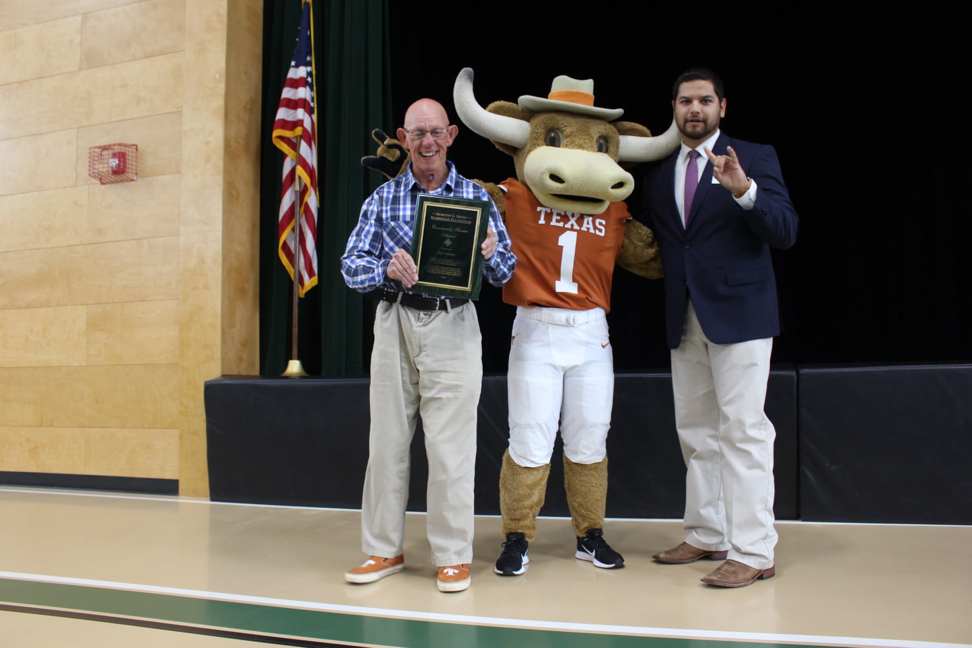 An man holding a plaque stands next to a person in a Texas Longhorns mascot costume and a man in a suit.