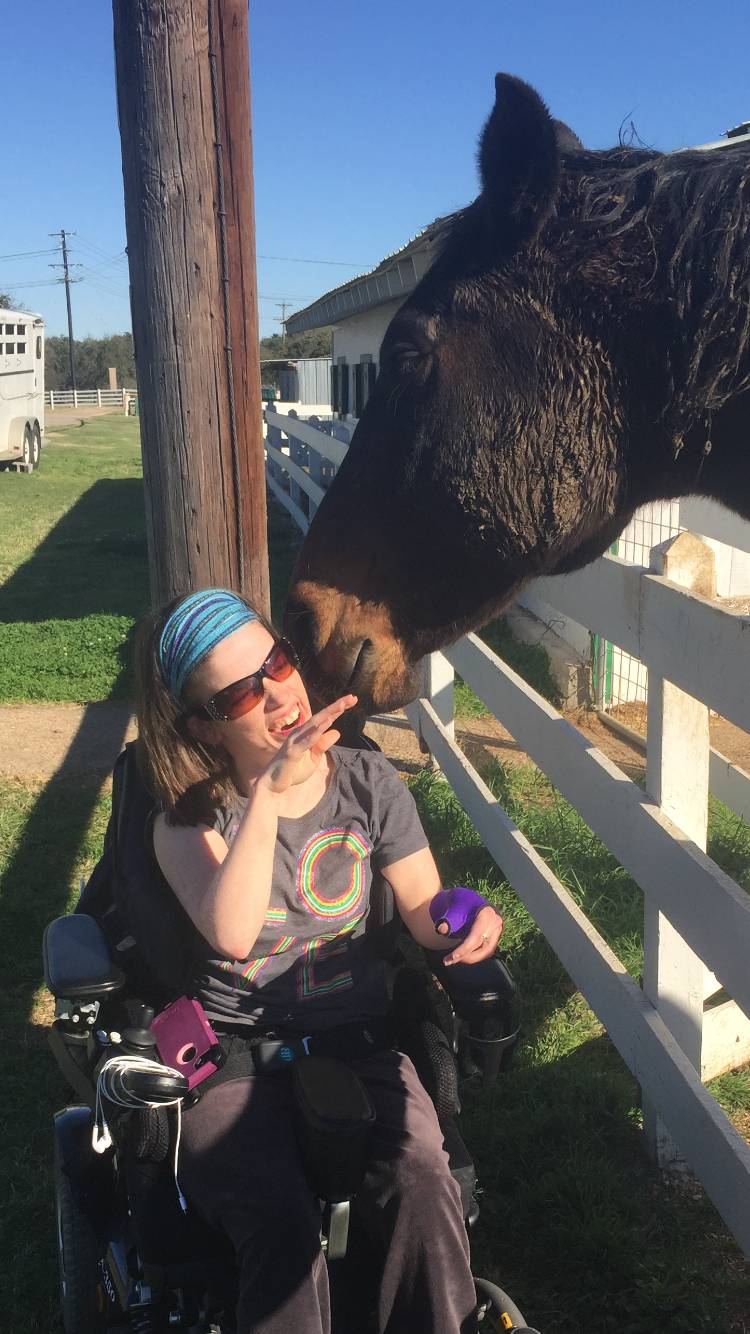 A resident in a power wheelchair smiles and pets a horse in a corral.