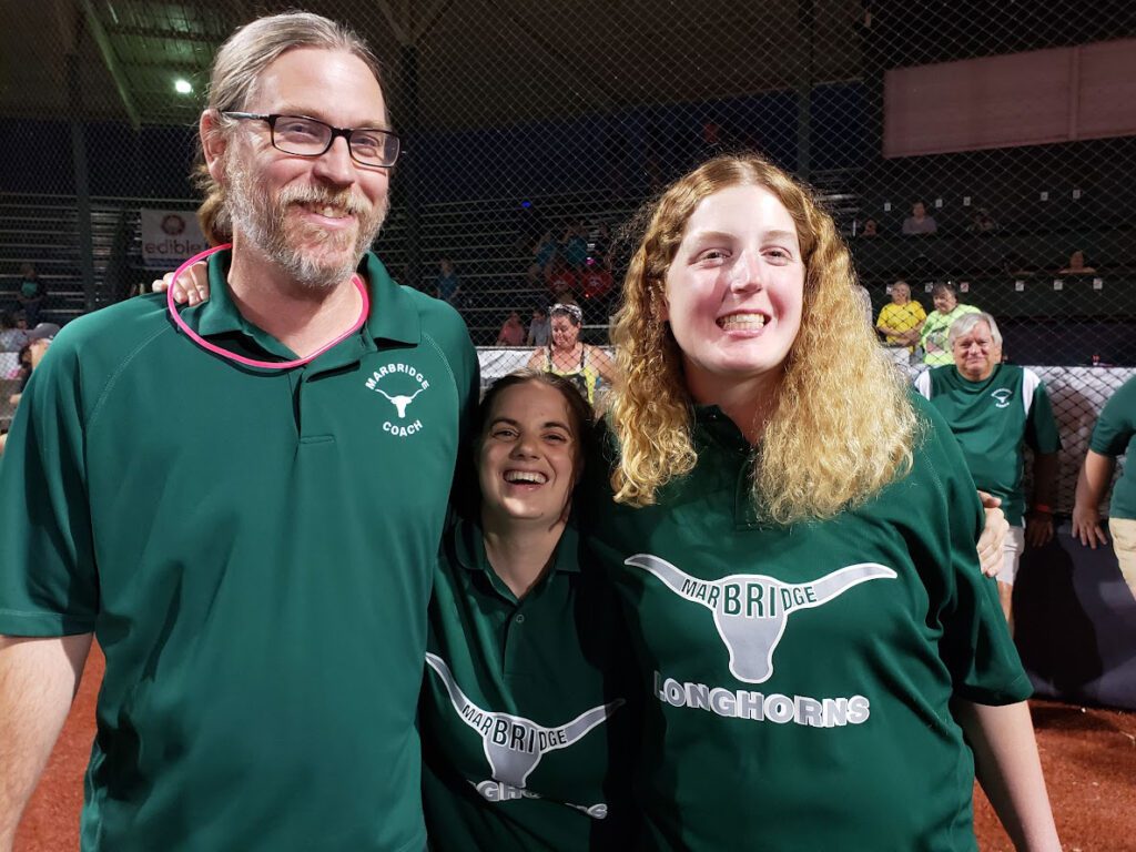Three people wearing green "Marbridge Longhorns" shirts stand smiling in a sports arena.